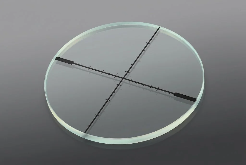 Circular glass reticle with crossed scales, tick marks and dark end blocks etched across the centre