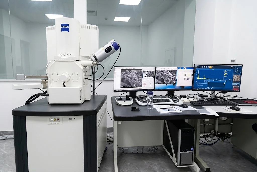 A ZEISS Gemini 1 scanning electron microscope beside a workstation with three monitors displaying microscopy images and measurement software in a clean metrology room